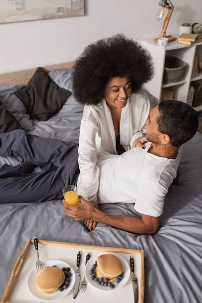 High angle view of african american woman smiling near young boyfriend and delicious pancakes with blueberries on bed — Stockfoto