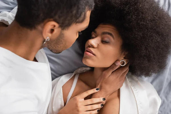 Vista de ángulo alto de hombre afroamericano cerca de novia seductora acostado en la cama con los ojos cerrados - foto de stock