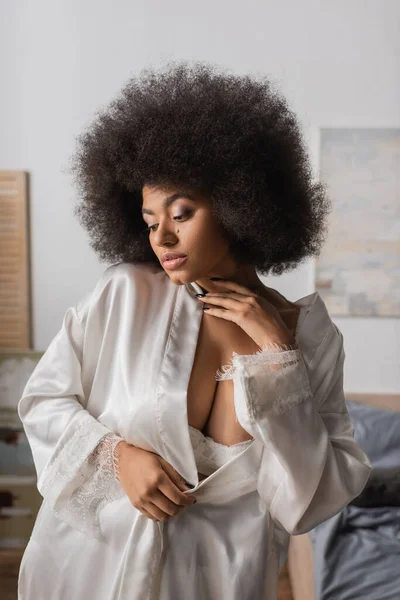 Sexy Afroamerikanerin in weißer Robe berührt Hals, während sie im Schlafzimmer posiert — Stockfoto