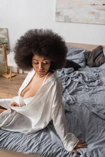 Seducente donna afroamericana in vestaglia bianca e reggiseno seduta su biancheria da letto grigia a casa — Foto stock