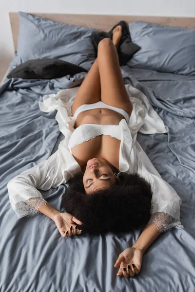Vista ad alto angolo della donna afro-americana in biancheria intima bianca sul corpo sexy sdraiato con gli occhi chiusi su biancheria da letto grigia — Foto stock