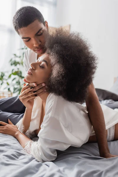 Uomo afroamericano toccando collo di donna bruna riccia in vestaglia di seta bianca sul letto a casa — Foto stock