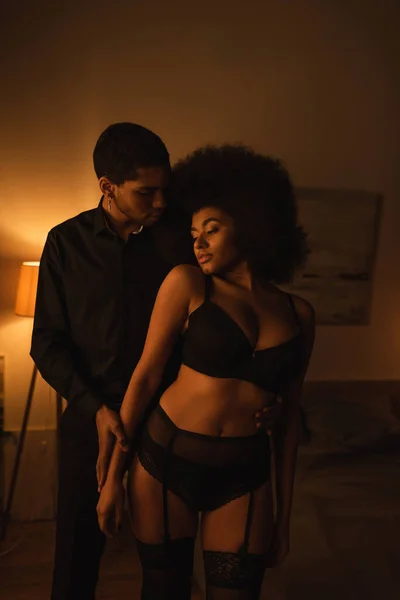 African american man in black shirt near passionate woman in sexy underwear in dark bedroom with lighting — Photo de stock