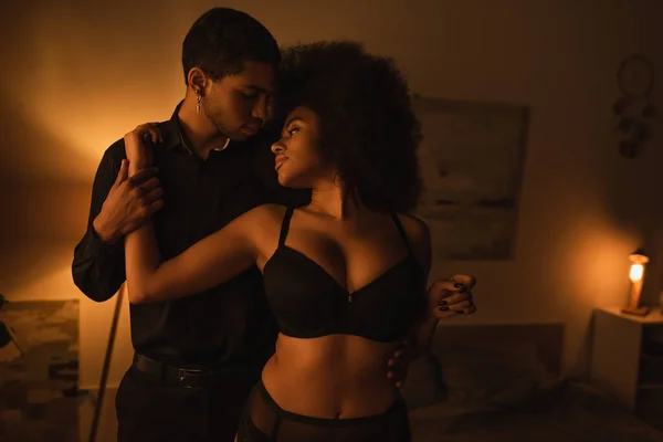 Sensual african american woman in black underwear hugging young boyfriend in dark room with luminous lamp — Photo de stock