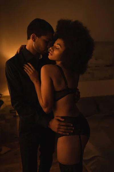 Seducente donna afroamericana in lingerie sexy che si abbraccia con il giovane fidanzato a casa di notte — Foto stock