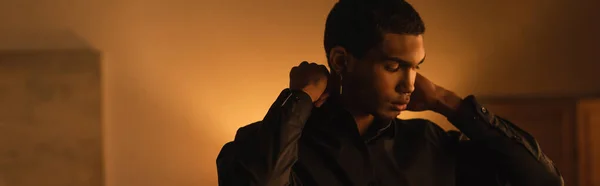 Молодий афроамериканський чоловік регулює комір чорної сорочки ввечері вдома, банер — стокове фото