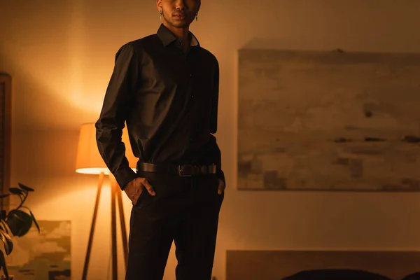 Cropped view of young african american man standing with hands in pockets of black trousers in dark room with lighting — Stock Photo