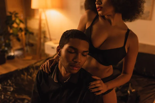 African american woman in black bra embracing shoulders of boyfriend sitting in bedroom with closed eyes — Stock Photo