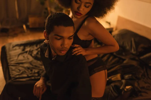 Sexy african american woman in underwear hugging shoulders of young man sitting on bed at night — Stock Photo