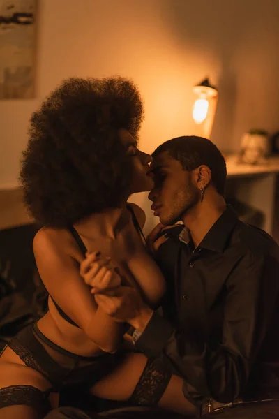 Young man in black shirt near seductive african american girlfriend sitting on bed in sexy underwear — Photo de stock