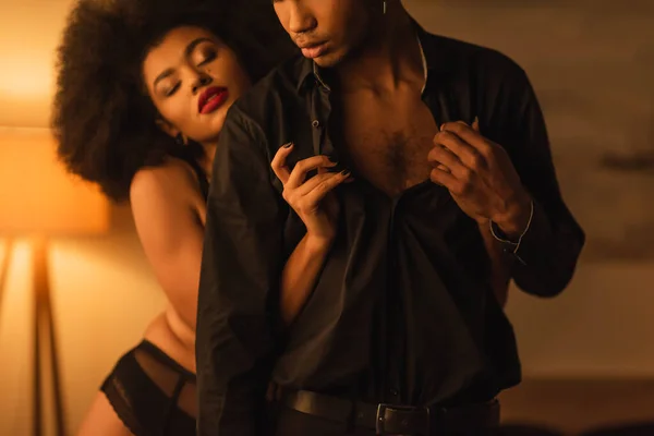 Sexy african american woman with closed eyes undressing young man in dark room with lighting — Stock Photo