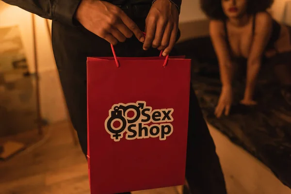 Vista recortada del hombre sosteniendo bolsa de papel de la tienda de sexo cerca de la novia afroamericana sobre fondo borroso - foto de stock