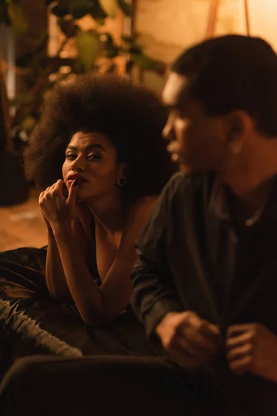 Curly african american woman looking at blurred boyfriend unbuttoning black shirt in bedroom — Fotografia de Stock