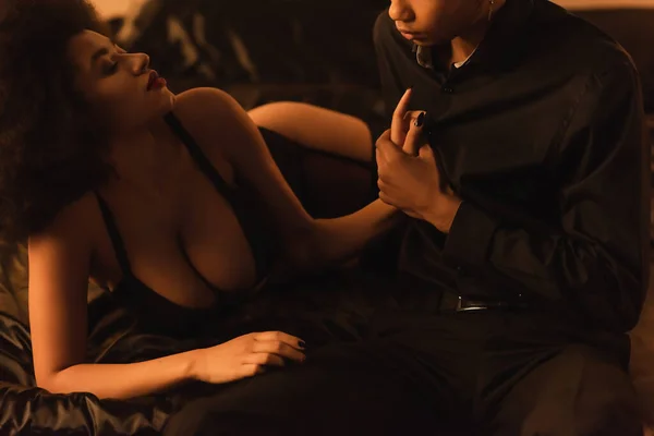 Young man holding hand of seductive african american woman on black silk bedding at home — Stock Photo