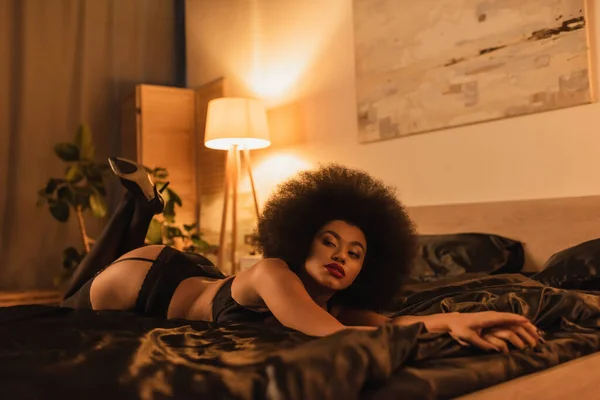 Сексуальна афроамериканська жінка, дивлячись далеко, лежачи в нижній білизні на чорному шовковій підстилці в спальні з яскравою лампою — стокове фото
