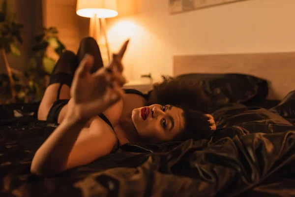 Appassionata donna afroamericana sdraiata su biancheria da letto in raso nero e guardando la fotocamera in primo piano sfocato — Foto stock