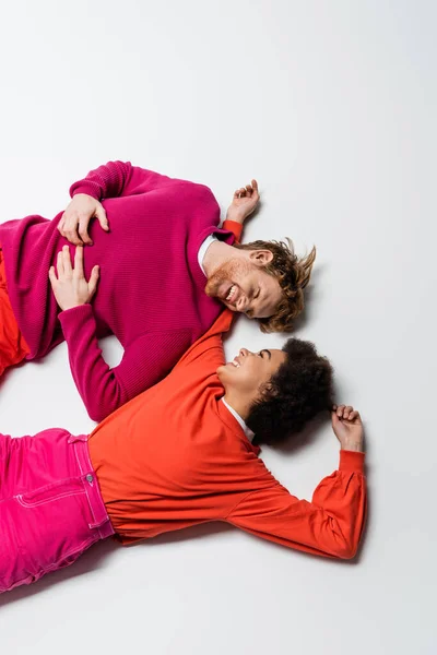 Top view of happy redhead man and curly african american woman in magenta color clothes lying on white — Stock Photo