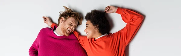 Top view of happy redhead man and curly african american woman in magenta color clothes lying on white, banner — Stock Photo