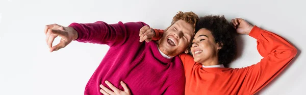 Vue du haut de l'homme rousse heureux dans des vêtements de couleur magenta pointant du doigt près de frisée femme afro-américaine sur blanc, bannière — Photo de stock