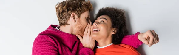 Top view of redhead man whispering in ear of cheerful african american woman lying on white, banner — Stock Photo