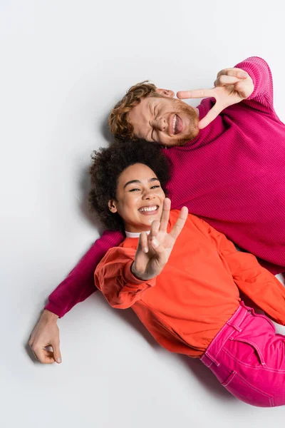 Вид зверху щасливого рудого чоловіка і кучерявої афроамериканської жінки в кольоровому одязі пурпурового кольору, що показує знак миру на білому — стокове фото