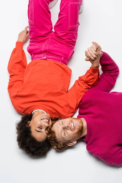 Top view of pleased redhead man and curly african american woman in magenta color clothes lying on white — Stock Photo