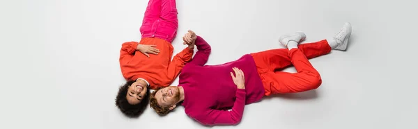 Top view of happy redhead man and joyful african american woman in magenta color clothes lying on white, banner — Photo de stock