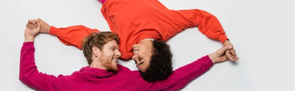Top view of happy redhead man in magenta color clothes and curly african american woman holding hands on white, banner — Stock Photo