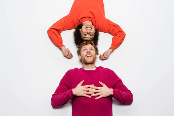 Top view of happy redhead man in magenta color clothes and excited african american woman lying on white — Photo de stock