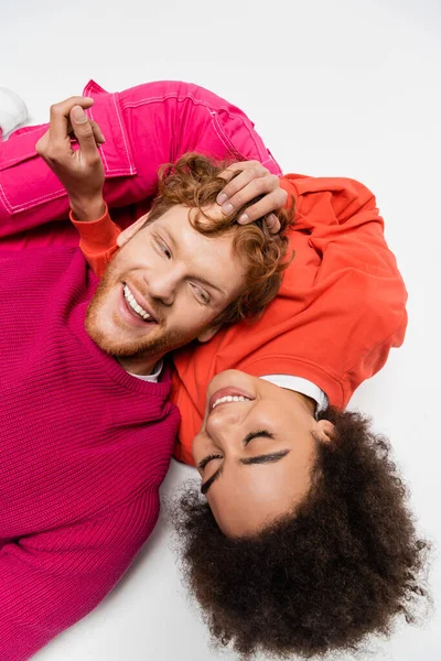 Top view of cheerful african american woman lying with redhead man in magenta color sweatshirt on white — Stock Photo