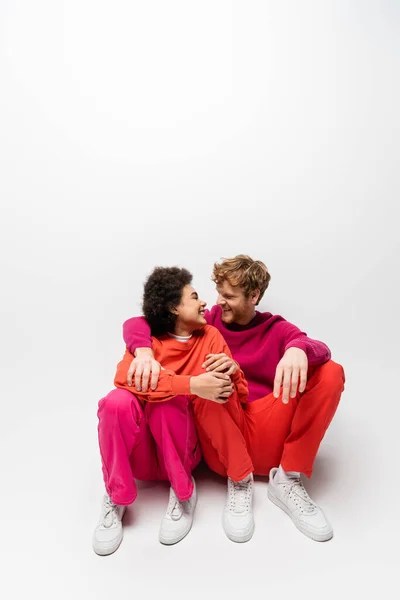 Positive african american woman sitting with happy man in magenta color sweatshirt on white — Stock Photo