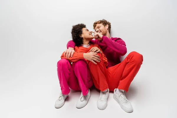 Positive man toughing nose of african american woman in magenta color clothes sitting on white — Stock Photo
