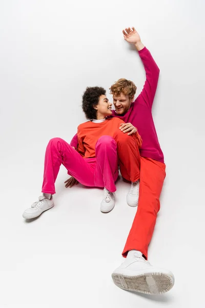 Positive african american woman sitting with happy man in magenta color clothes raising hand on white — Fotografia de Stock
