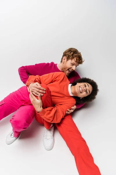 Overhead view of happy redhead man in magenta color sweatshirt hugging cheerful african american woman on white — Stock Photo