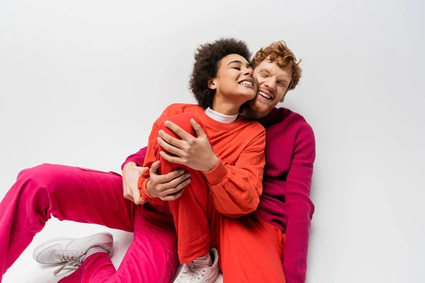 Happy redhead man in magenta color sweatshirt hugging smiling african american woman on white — Stock Photo