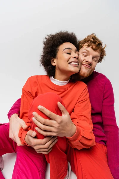 Happy redhead man in magenta color sweatshirt hugging cheerful african american woman on grey — Photo de stock