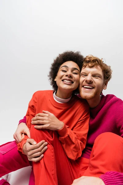 Positive redhead man in magenta color sweatshirt hugging cheerful african american woman on grey — Stock Photo
