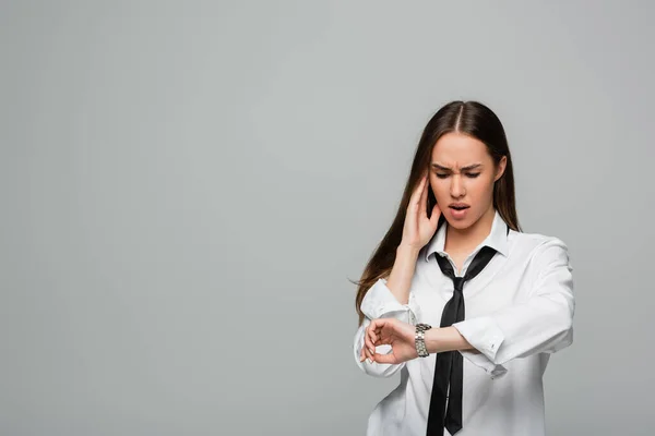 Shocked and brunette woman in white shirt and tie looking at wristwatch isolated on grey — Stock Photo
