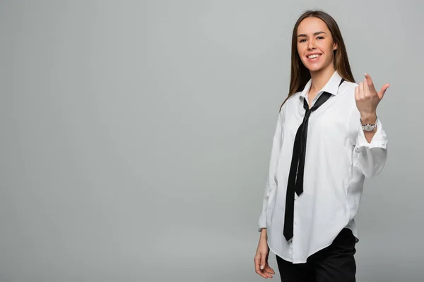 Cheerful young woman in white shirt and tie looking at camera while pointing with finger isolated on grey — Stock Photo
