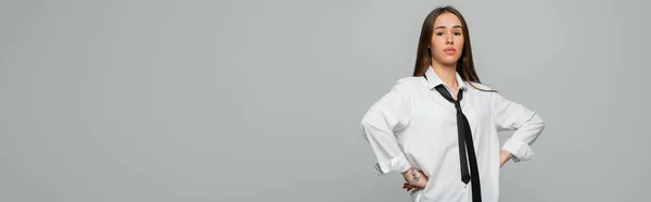 Confident young woman in white shirt and tie looking at camera while standing with hands on hips isolated on grey, banner — Stock Photo