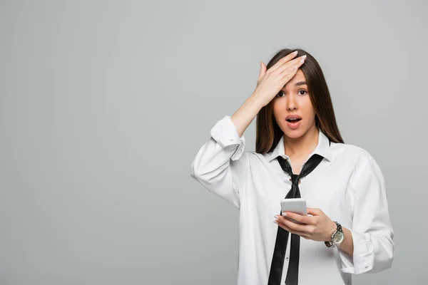 Шокированная молодая женщина в белой рубашке и галстуке держа смартфон изолирован на сером — стоковое фото