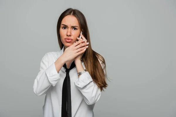 Displeased young woman in white shirt and tie talking on smartphone isolated on grey — Stock Photo