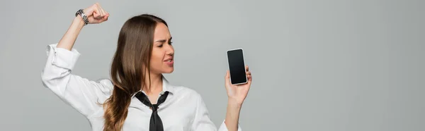 Young woman in shirt and tie standing with clenched fist and holding smartphone with blank screen isolated on grey, banner — Stock Photo