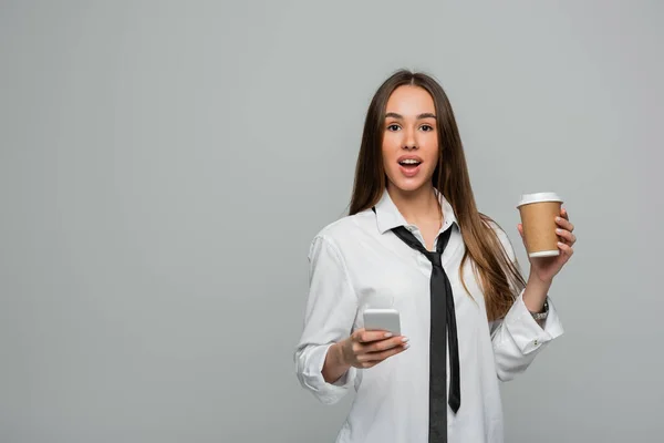 Amazed young woman in white shirt with tie holding smartphone and paper cup with coffee to go isolated on grey — Stock Photo