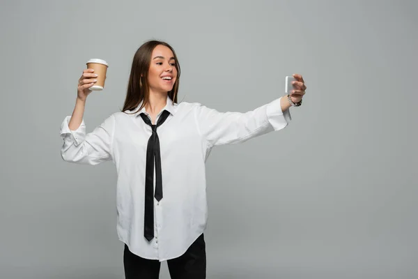 Happy young woman in white shirt with tie taking selfie with paper cup isolated on grey — Stock Photo