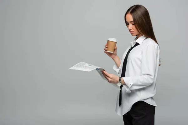 Young woman in white shirt with tie reading newspaper while holding paper cup isolated on grey — Stock Photo