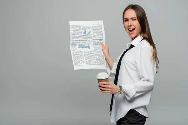 Amazed young woman in white shirt with tie holding newspaper and paper cup isolated on grey — Stock Photo