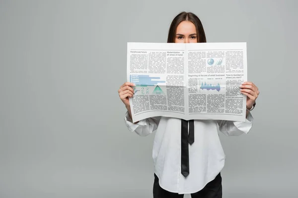 Young woman in white shirt with tie covering face with newspaper isolated on grey — Photo de stock