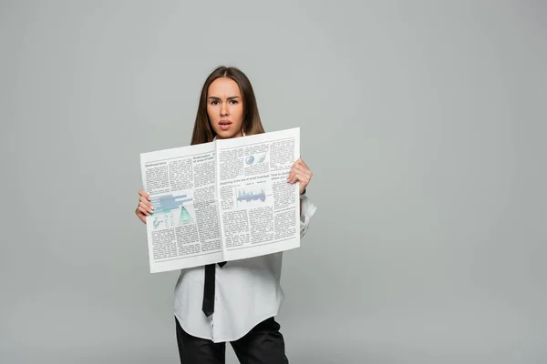 Shocked young woman in white shirt with tie holding newspaper while looking at camera isolated on grey — Stock Photo