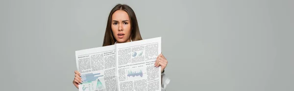 Shocked young woman holding newspaper while looking at camera isolated on grey, banner — Stock Photo
